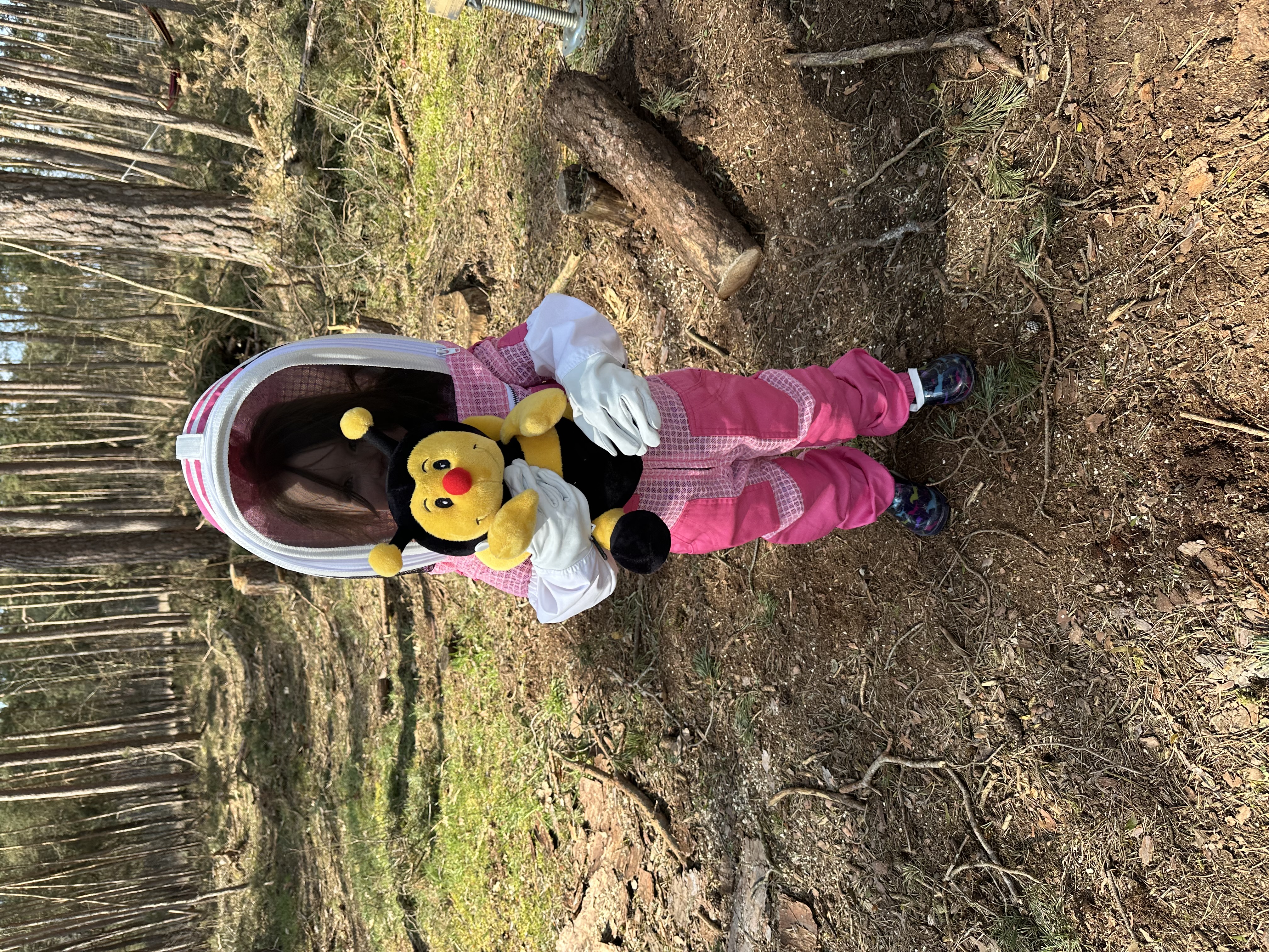 Kleine Imkerin mit Schutzanzug und Bienen-Kuscheltier am Bienenstand